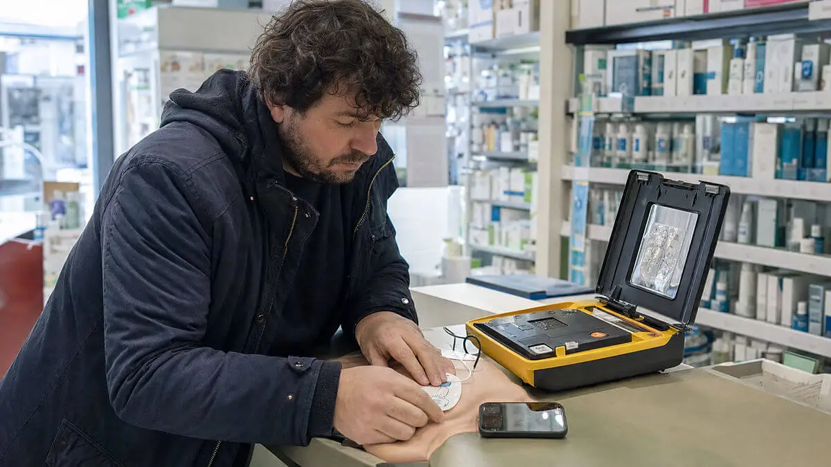 Interior de farmacia urbana; mostrador con DEA abierto y parches visibles; un transeúnte coloca parches sobre una superficie