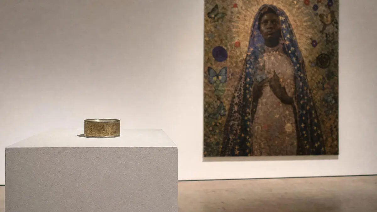 Bodegón conceptual de una lata metálica sellada sobre un pedestal de galería y, al fondo, una obra religiosa contemporánea co